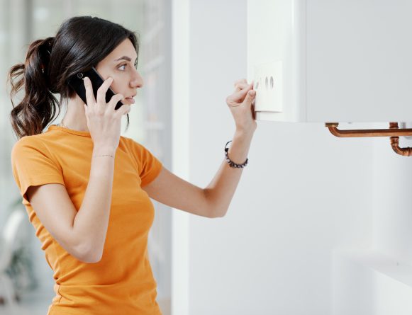 Puzzled woman fiddles with a combi boiler while on the phone to a gas and heating engineer